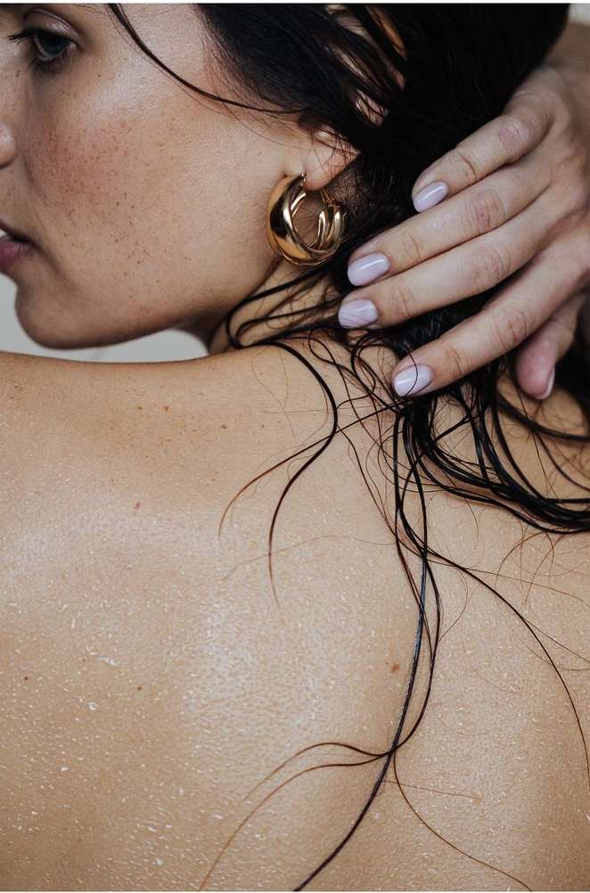 Close-up portrait of a woman with wet dark hair touching her ear, wearing a gold hoop earring, photographed by Lucie Sassiat