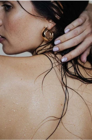 Close-up portrait of a woman with wet dark hair touching her ear, wearing a gold hoop earring, photographed by Lucie Sassiat