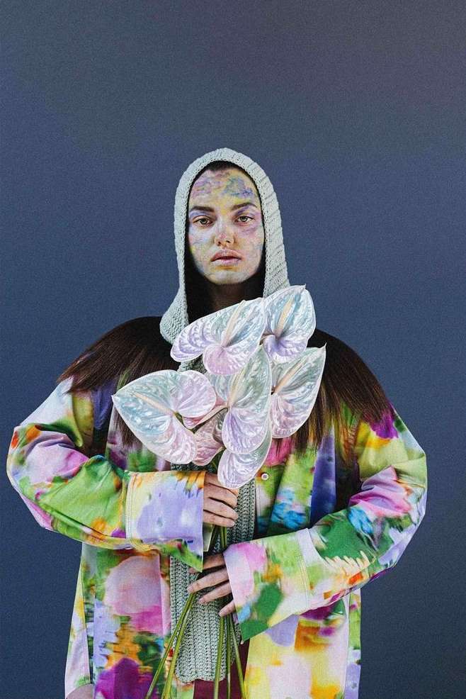 Artistic portrait of a woman with face paint wearing a knitted hood and a patterned coat, holding a bouquet of anthurium flowers.