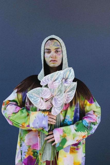 Artistic portrait of a woman with face paint wearing a knitted hood and a patterned coat, holding a bouquet of anthurium flowers.