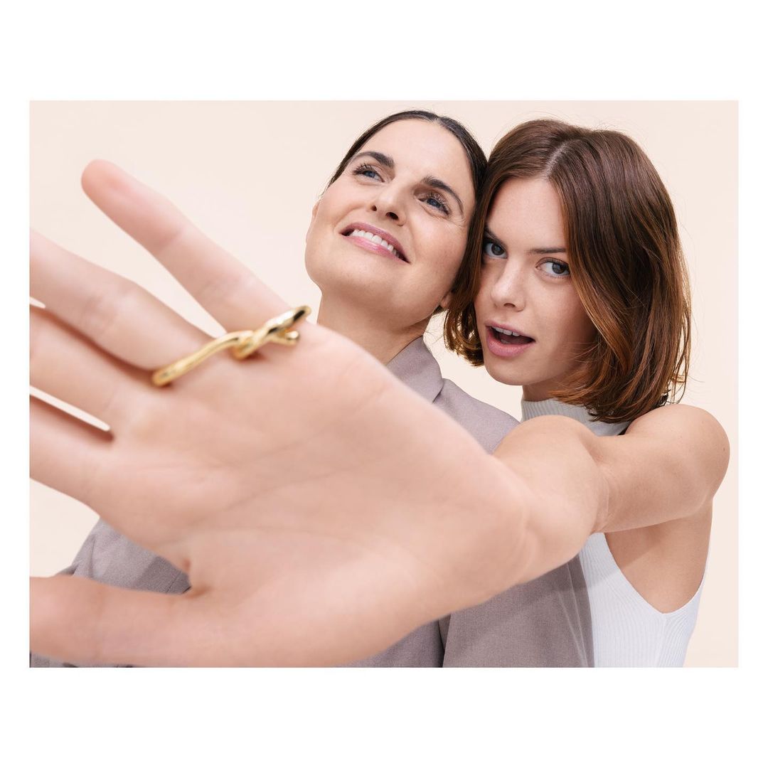 Two women posing closely with a gold ring worn on an outstretched hand against a plain neutral background.