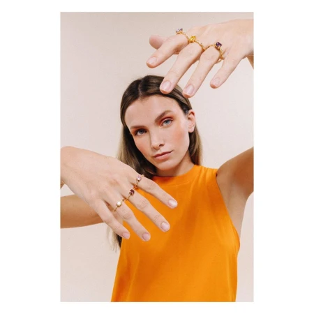 Woman in orange top holding her hands up to display gemstone rings on multiple fingers against a plain background