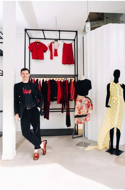 Man in a black suit stands in a clothing store next to two mannequins, with a clothing rack full of red clothes in the background.