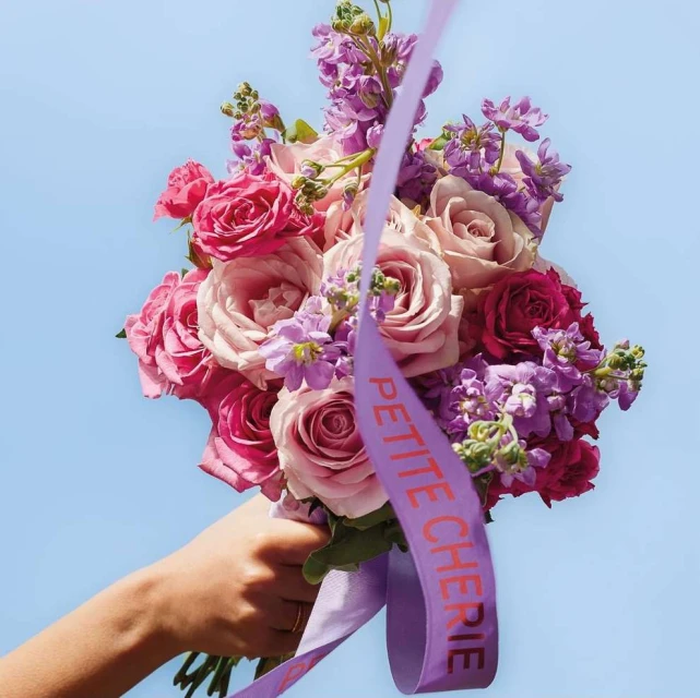 A hand holds a vibrant bouquet of pink and purple roses against a bright blue sky, accented by a ribbon with the words 'Petite Cherie'.