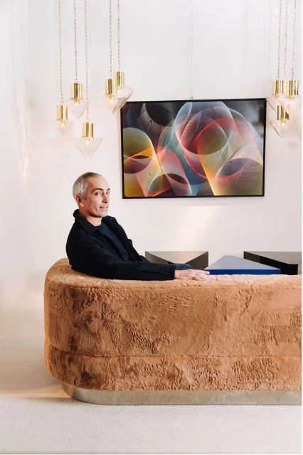 Interior shot featuring a man seated behind a textured desk, illuminated by hanging lights, with abstract art on the wall. 