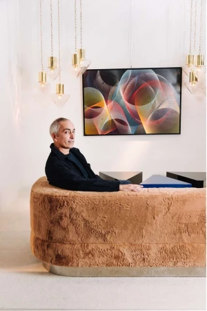 Interior shot featuring a man seated behind a textured desk, illuminated by hanging lights, with abstract art on the wall. 