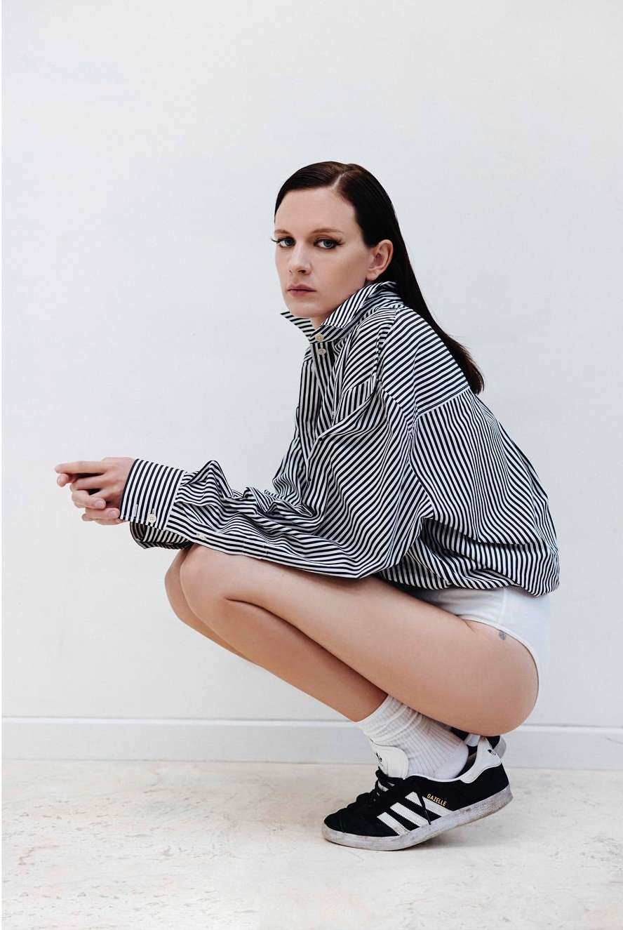 A woman squats indoors wearing a striped shirt, socks, and sneakers, striking a candid pose against a bright white wall.