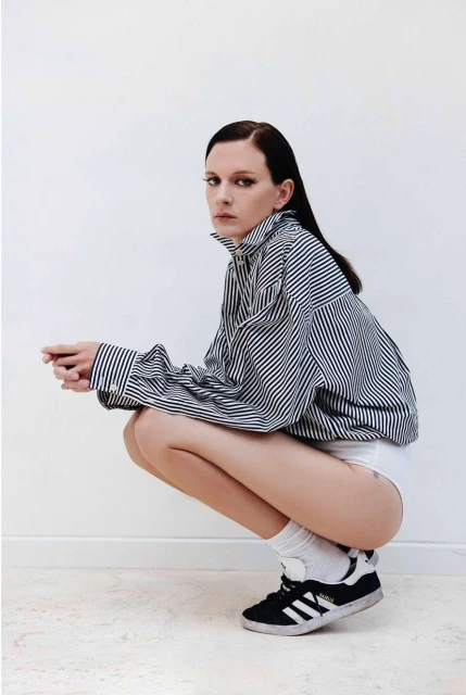 A woman squats indoors wearing a striped shirt, socks, and sneakers, striking a candid pose against a bright white wall.