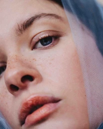 Close-up portrait of a woman with freckles and blue eyes wearing a sheer blue veil by photographer Lucie Sassiat.