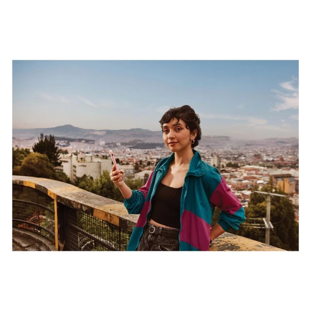 Young woman poses for a selfie in front of an urban cityscape. She holds up a phone, wearing a colorful jacket, with a city backdrop.
