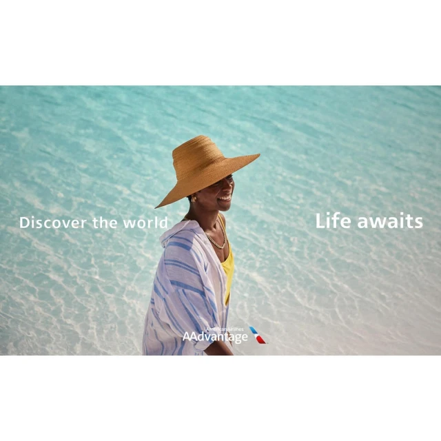 Smiling woman in a straw hat and striped cover-up walks along a tropical beach with turquoise water, promoting world travel.