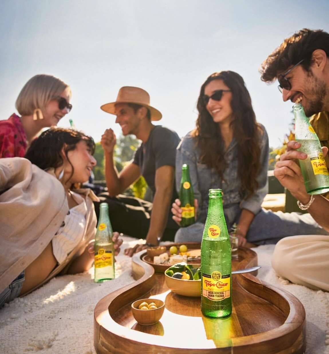 Group of five diverse young adults outside enjoying sparkling water and snacks on a guitar-shaped tray, socializing, and smiling in the sun.