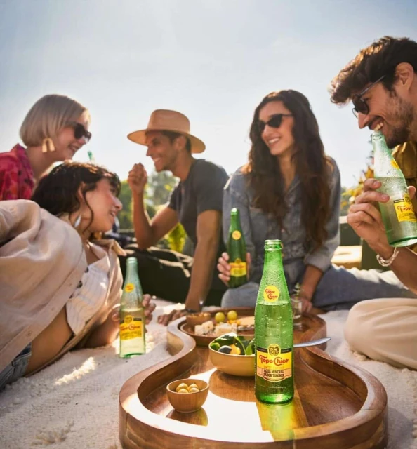 Group of five diverse young adults outside enjoying sparkling water and snacks on a guitar-shaped tray, socializing, and smiling in the sun.