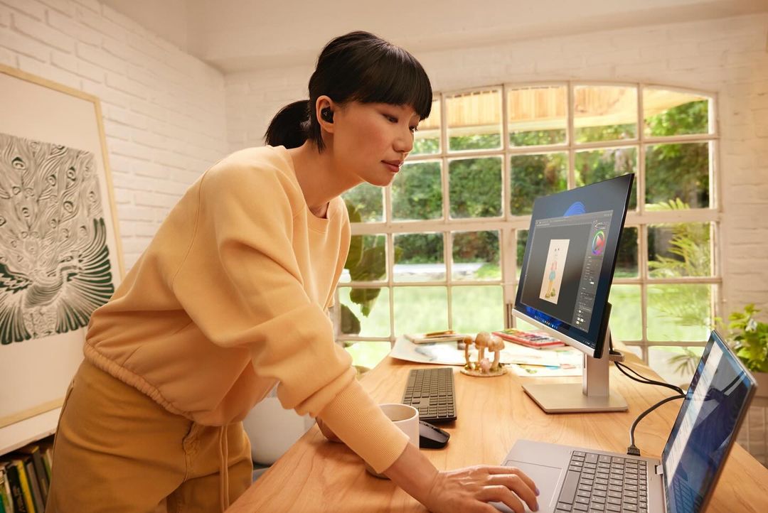 Woman works on a laptop and monitor in a home office setting, wearing wireless earbuds and a yellow sweatshirt, with natural light.