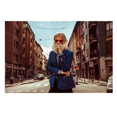 Stylish elderly man with a beard and red glasses stands on a city street with tall buildings and parked cars on either side.