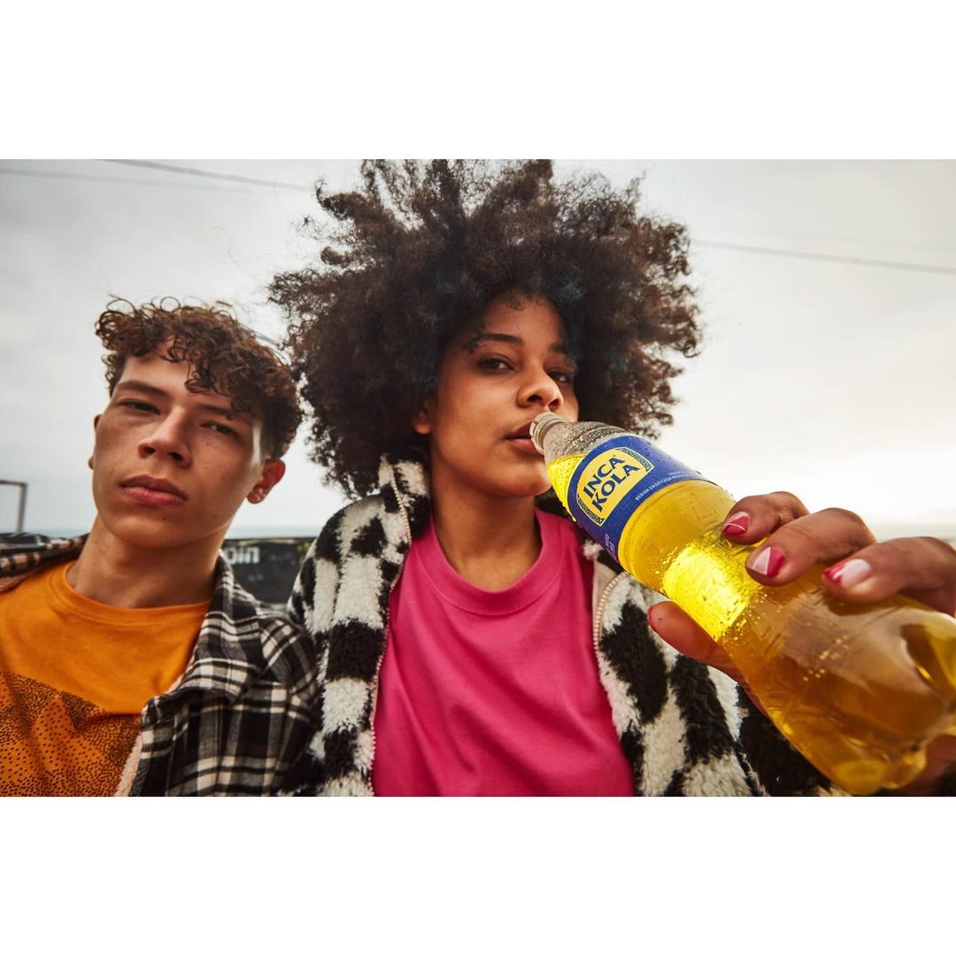 Two young adults pose with an Inca Kola bottle outdoors; one drinks while the other looks at the camera, dressed in trendy streetwear.