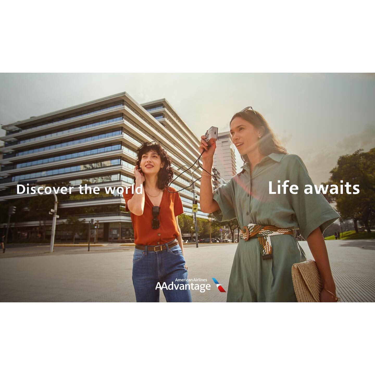 Two women in casual outfits are walking in an urban setting, with one taking a photo of a modern building on a sunny day.