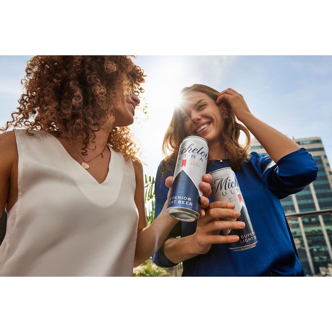 Two women smiling and holding Michelob Ultra beer cans outdoors on a sunny day with modern architecture in the background.