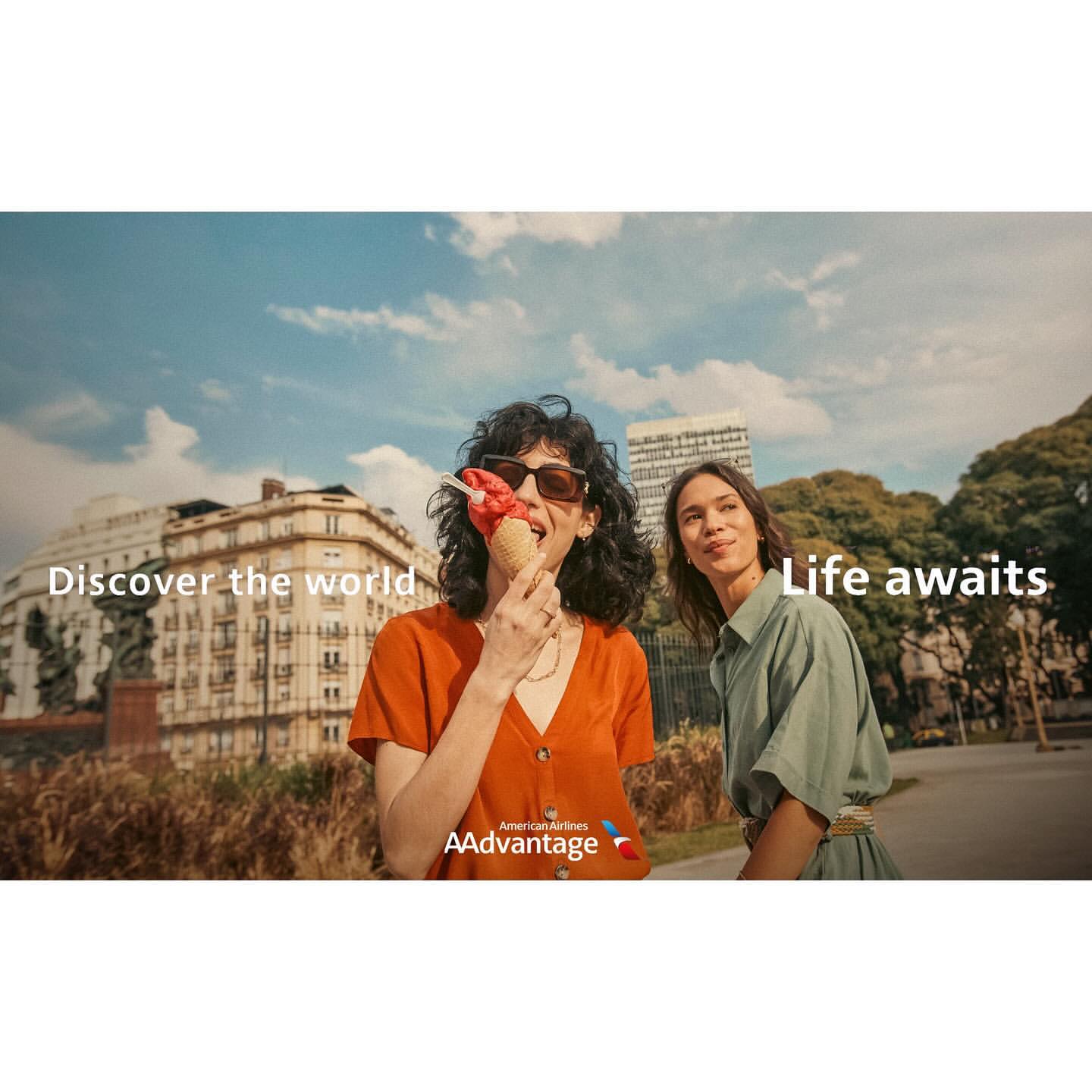 Two women enjoy ice cream and city views, wearing sunglasses and casual summer clothes, with blue sky and urban architecture in the background.