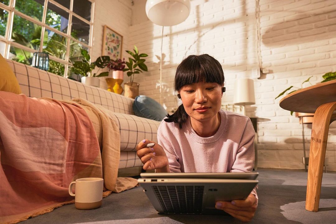 Young Asian woman using a laptop on the floor of a living room with a sofa, table, and plants, creating a cozy and productive workspace.