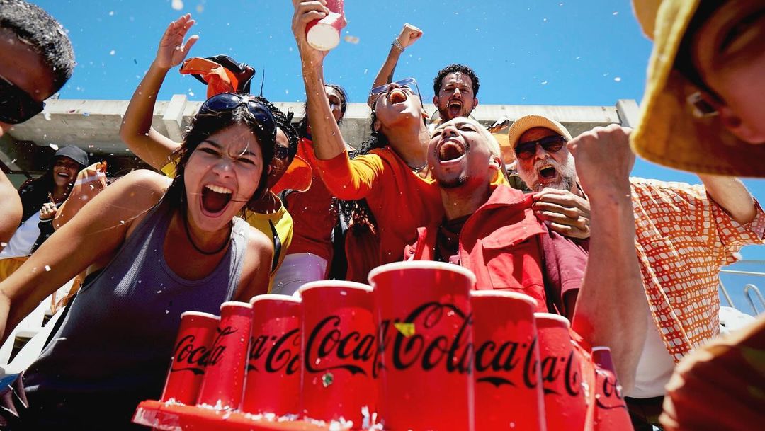 Enthusiastic group celebrates outdoors with Coca-Cola cups; confetti showers down on diverse adults cheering and laughing under a clear sky.