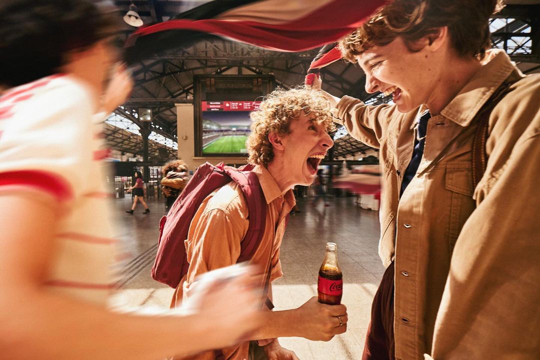 Excited group of young adults celebrates while watching a soccer game at a train station, holding a Coca-Cola bottle, indoors.