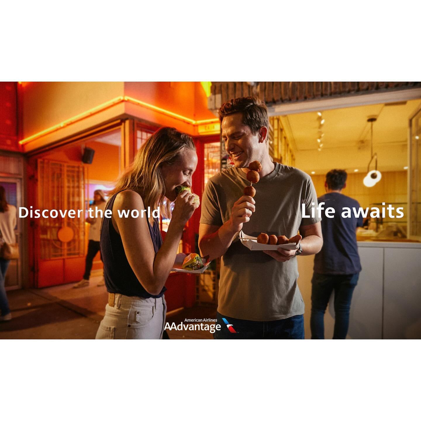 Couple enjoys street food at an outdoor eatery with glowing lights; American Airlines AAdvantage logo visible, promoting travel and culinary experiences.