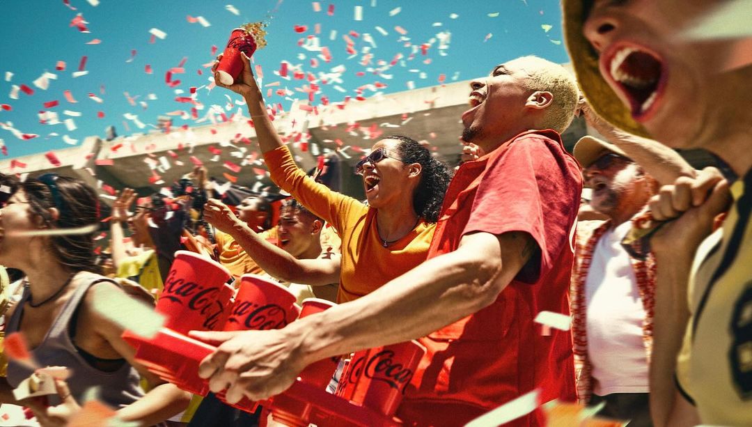 Excited crowd celebrates with red Coca-Cola cups and confetti under a sunny sky, showcasing youthful energy and brand integration.