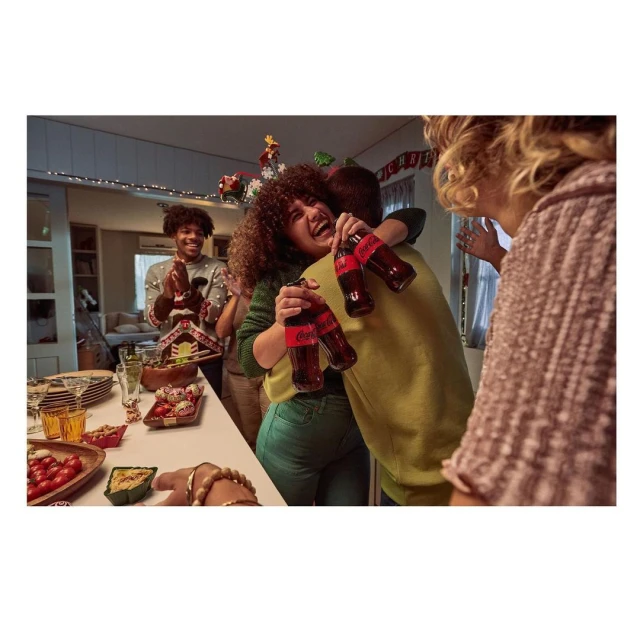 A happy group of people celebrate indoors, hugging and laughing while holding Coca-Cola bottles, with holiday decorations and snacks.