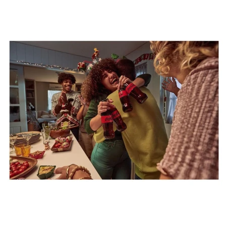 A happy group of people celebrate indoors, hugging and laughing while holding Coca-Cola bottles, with holiday decorations and snacks.