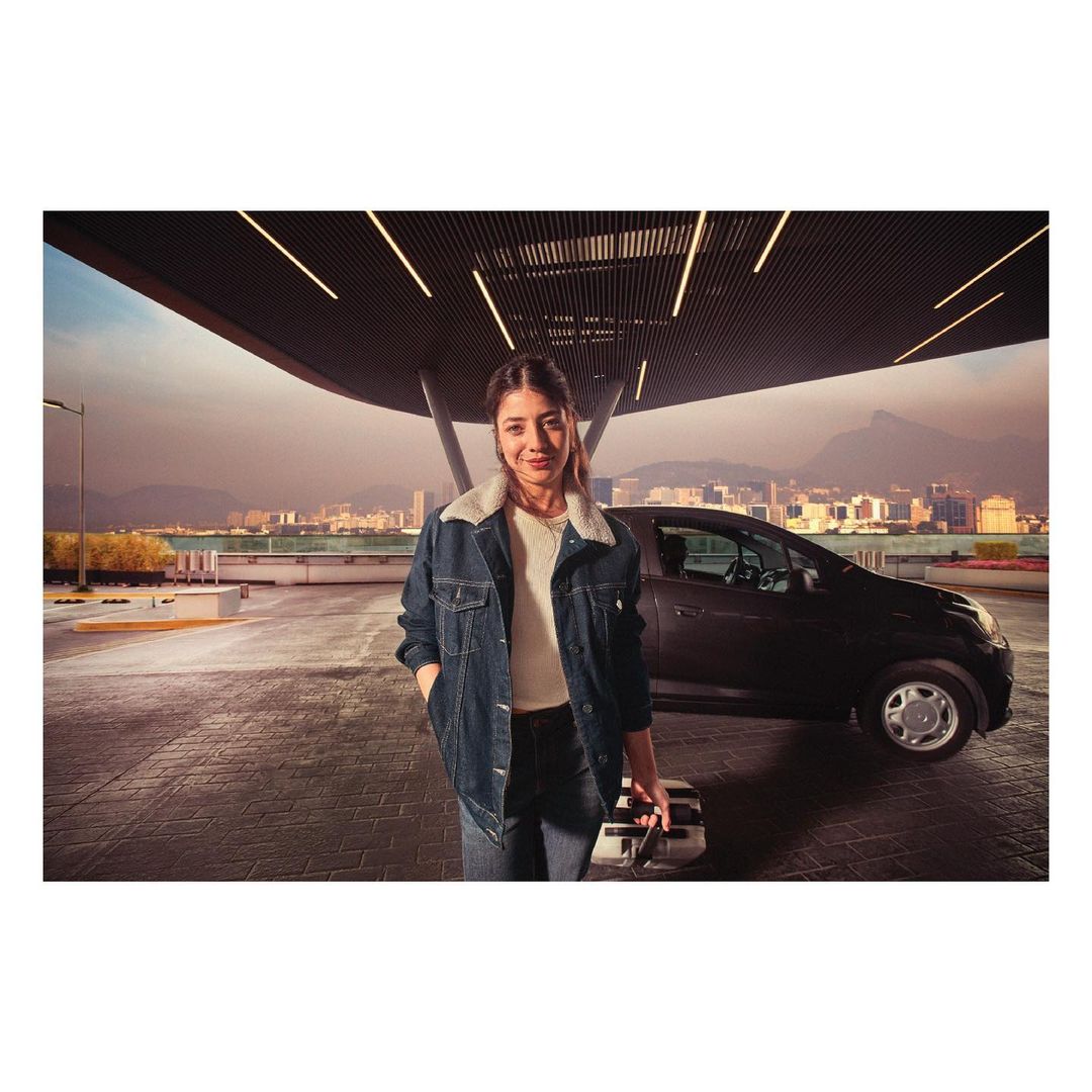 A woman in a denim jacket stands near a compact car in an urban setting, holding a suitcase, with a cityscape in the background.