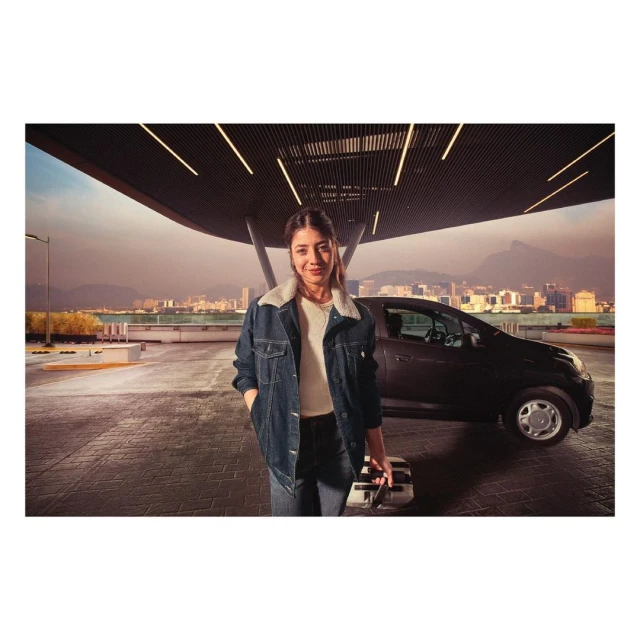 A woman in a denim jacket stands near a compact car in an urban setting, holding a suitcase, with a cityscape in the background.