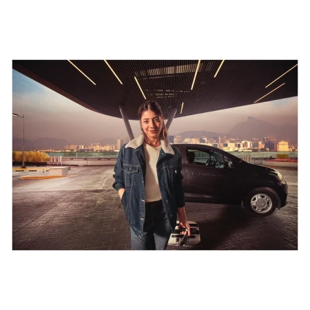 A woman in a denim jacket stands near a compact car in an urban setting, holding a suitcase, with a cityscape in the background.