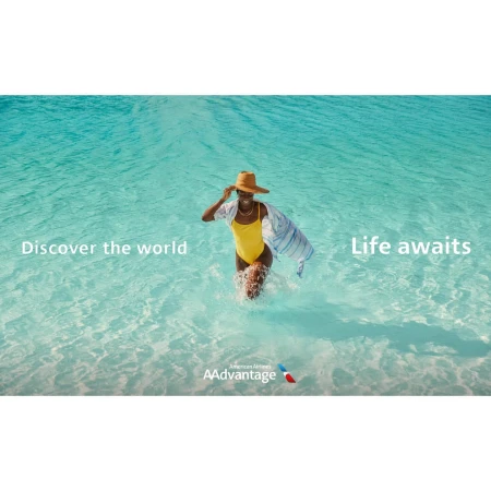 Woman in a yellow swimsuit wading in turquoise ocean water with a straw hat and striped cover-up, promoting travel.