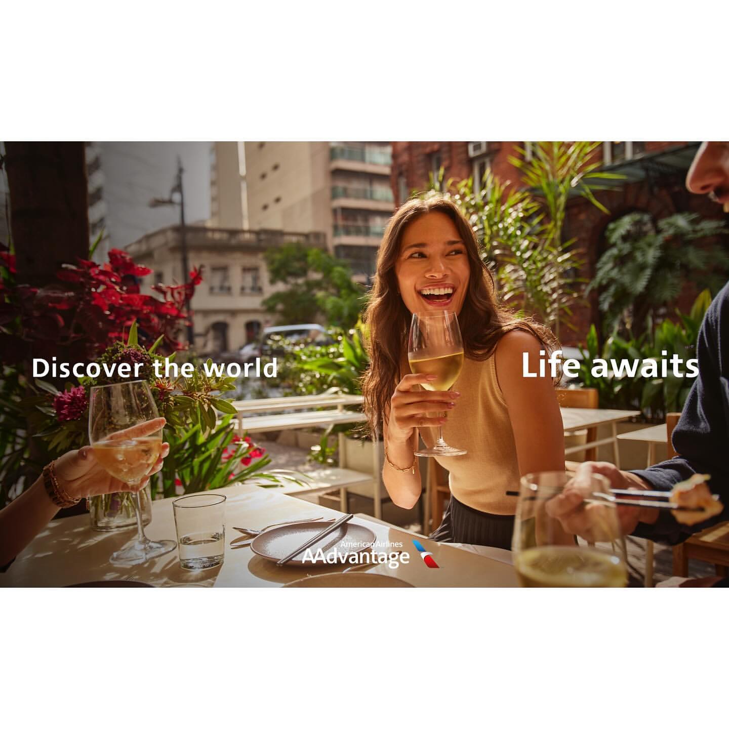A group of people sit at an outdoor table, sharing wine and food in a city setting, enjoying a meal with urban views.