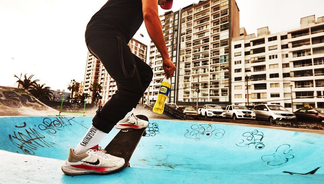 A person skateboarding in an urban skatepark, holding an Inca Kola bottle, with buildings in the background on a bright day.