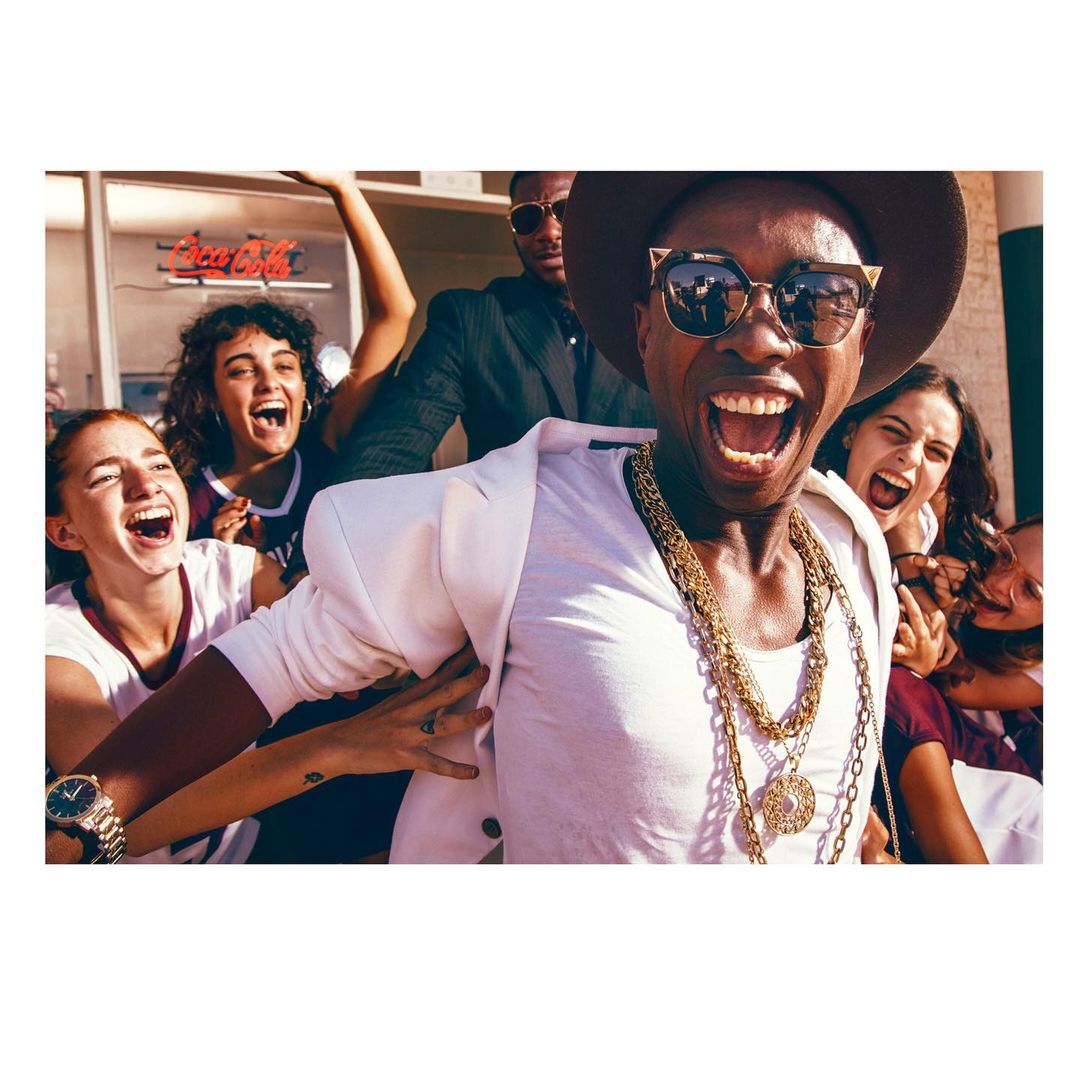 An excited group gathers around a stylish man in sunglasses, a white blazer, and gold chains, creating a lively, candid outdoor scene.