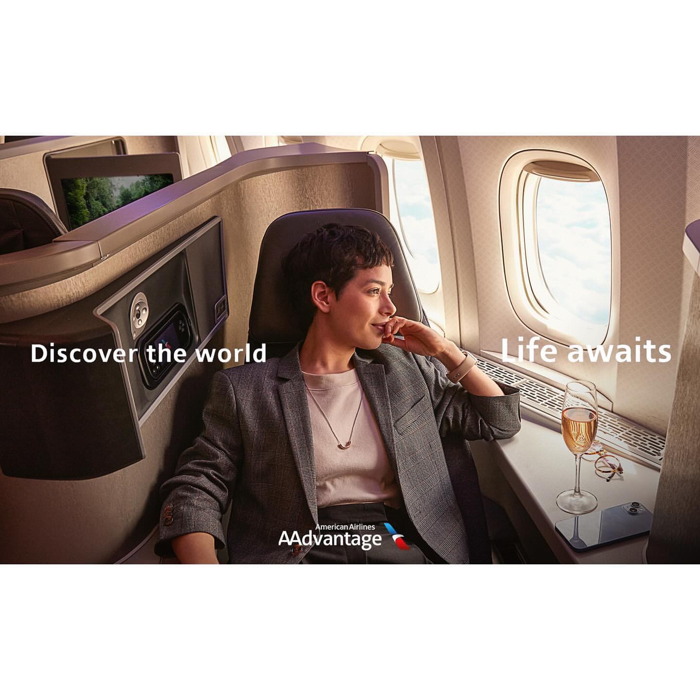 Adult female sits in first-class airplane seat with champagne, dressed in grey blazer and dark pants, looking out the window during flight.