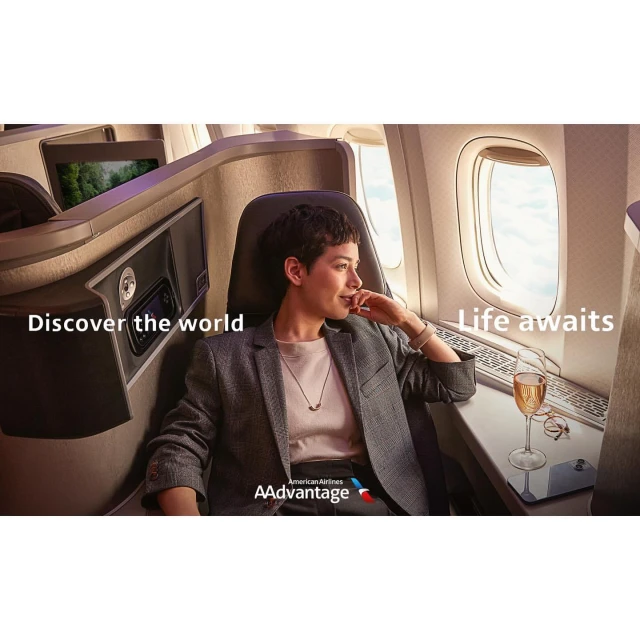 Adult female sits in first-class airplane seat with champagne, dressed in grey blazer and dark pants, looking out the window during flight.