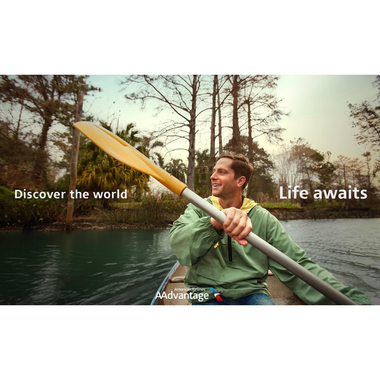 A smiling man paddles a canoe through a river surrounded by trees under the bright sky while enjoying the scenery.