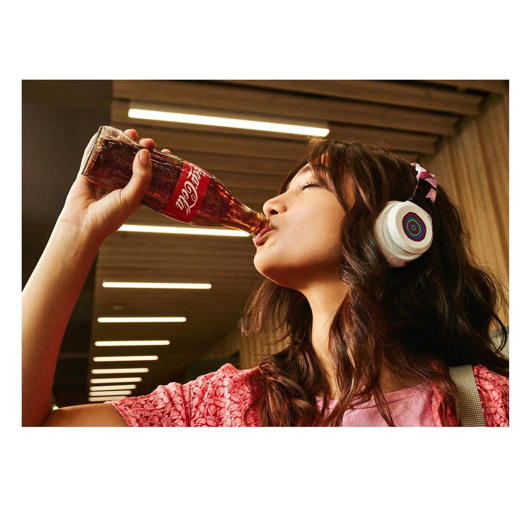Brunette woman drinks from a glass Coca-Cola bottle, wearing white headphones with colorful details in an indoor setting with warm lighting.