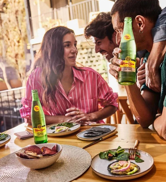 Three people at an outdoor table drink Topo Chico mineral water with tostadas. A woman in a pink shirt talks with two men outside.