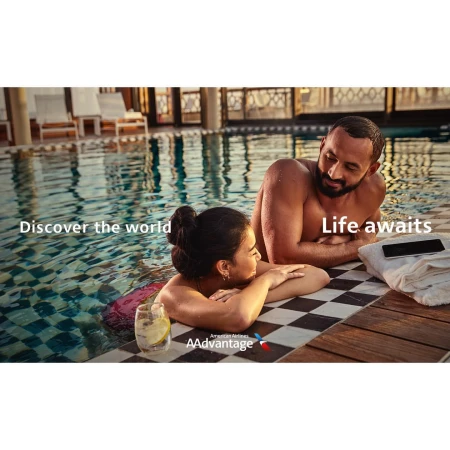 A man and a woman relaxing in an outdoor swimming pool resting their arms on the edge by a checkered patio deck.