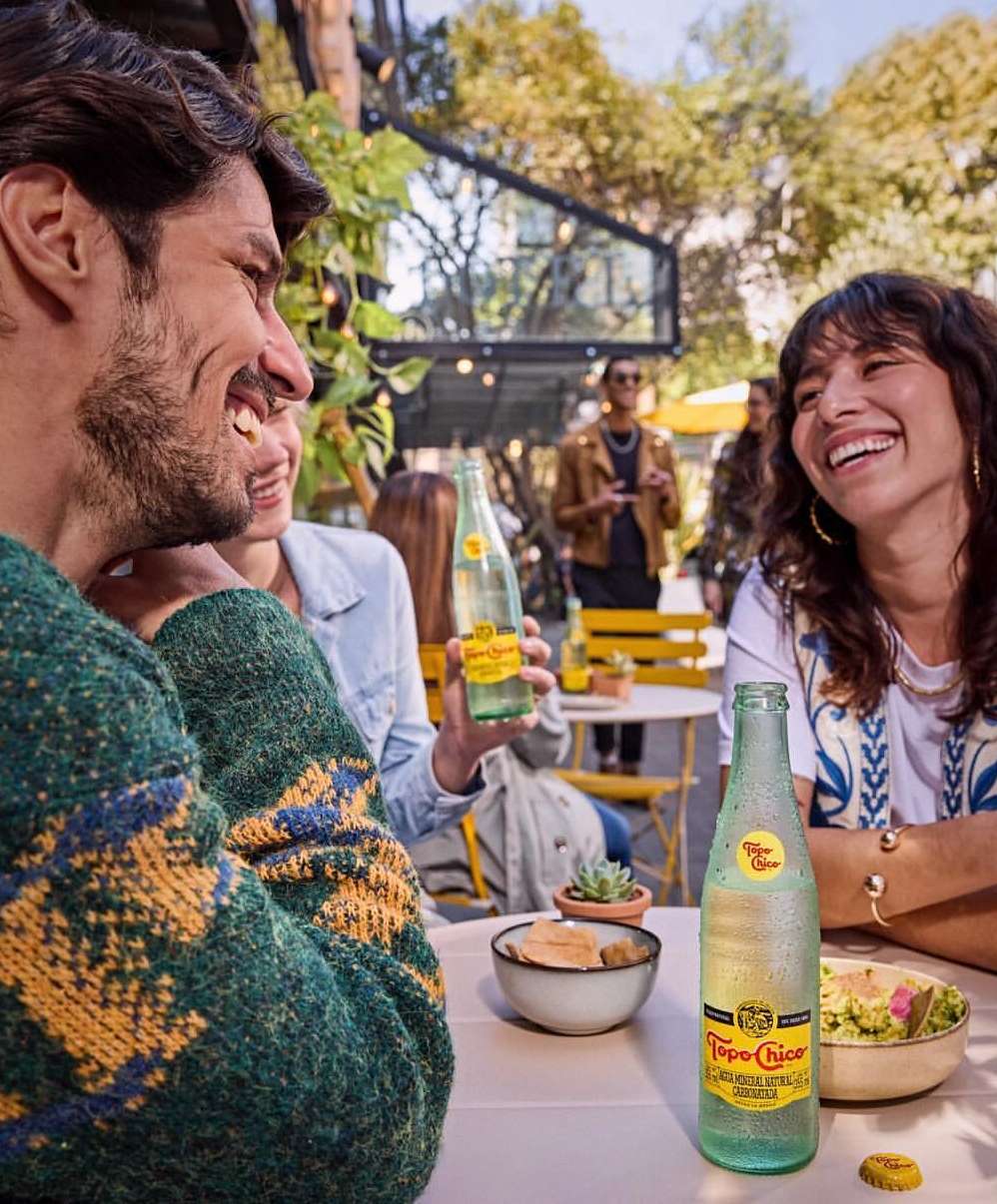A group of friends are seated at an outdoor cafe, laughing and enjoying Topo Chico mineral water on a sunny day with lush greenery.