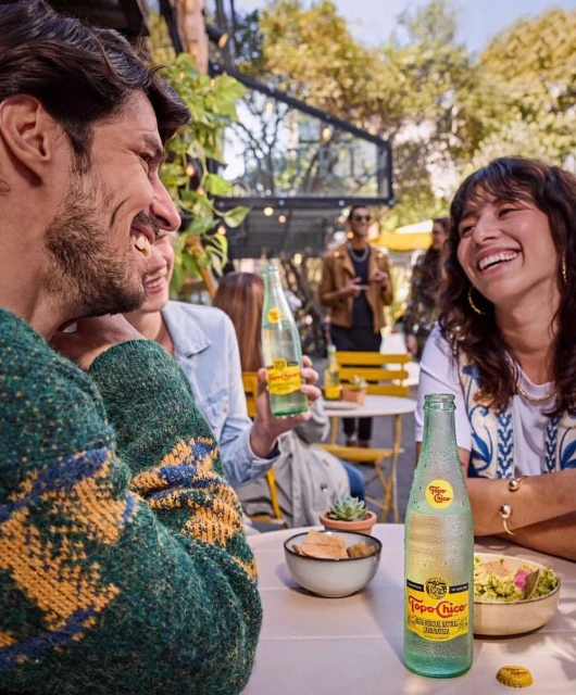 A group of friends are seated at an outdoor cafe, laughing and enjoying Topo Chico mineral water on a sunny day with lush greenery.