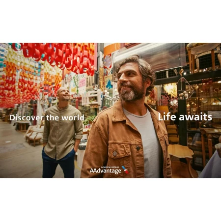 Commercial shot of two adult men in a vibrant market setting with hanging colorful decorations, and the American Airlines AAdvantage logo.