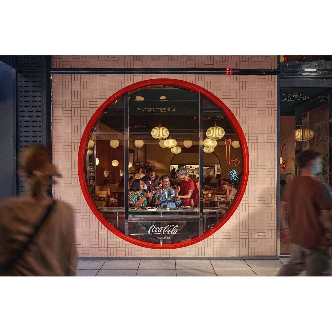 Patrons are seen through a large, round window, dining inside a restaurant with globe pendant lights and servers attending to the customers.