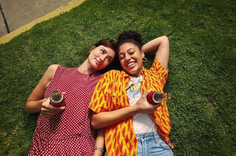 Two smiling women, laying on grass, holding glass soda bottles, wearing summer clothes, bright sunlight, joyful mood.