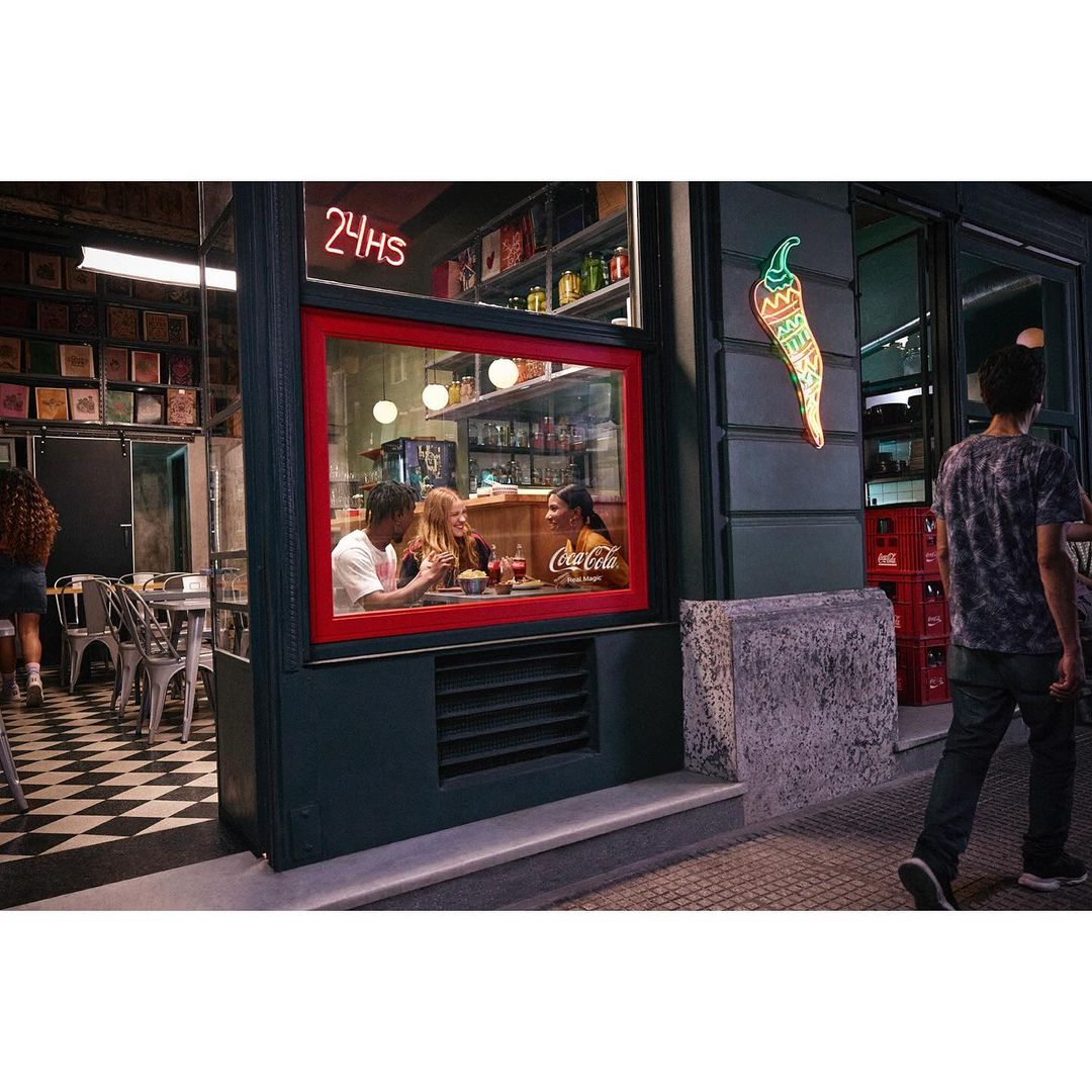 Three people sit inside a restaurant behind a window. Neon sign, chili pepper light, person walking by on sidewalk at night.