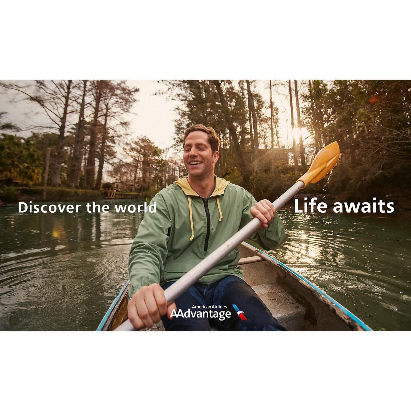 Man smiling in a canoe on a river surrounded by trees, holding a paddle; promotional image for American Airlines AAdvantage program.